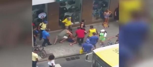 Na imagem um homem de camisa verde está sentado em cima das pernas do jovem imobilizando-o, enquanto outras pessoas assistem à cena. Na imagem um homem de camisa verde está sentado em cima das pernas do jovem imobilizando-o, enquanto outras pessoas assistem à cena.