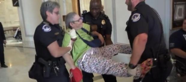 Disabled protester carried away from front of Mitch McConnel's office / Image screenshot CBS News via YouTube:https://youtu.be/qX_olozjUU8 Disabled protester carried away from front of Mitch McConnel's office / Image screenshot CBS News via YouTube:https://youtu.be/qX_olozjUU8