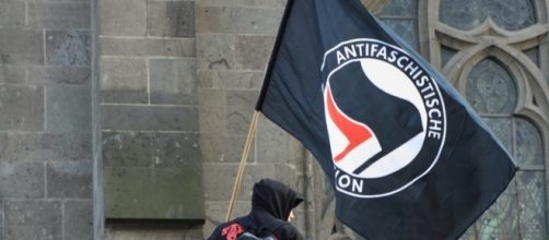 Antifa flag being held up before the Cologne Cathedral / [Image by Spaztacular via Flickr, CC BY 2.0]