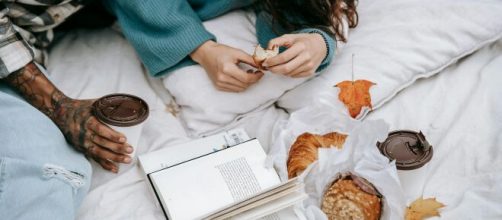 Ragazza che mangia un croissant sul letto con libro- © Pexels