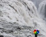 Una ragazza con un ombrello arcobaleno che osserva una cascata (©Pixabay)