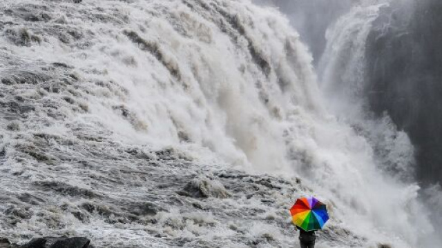 Una ragazza con un ombrello arcobaleno che osserva una cascata (&copy;Pixabay)